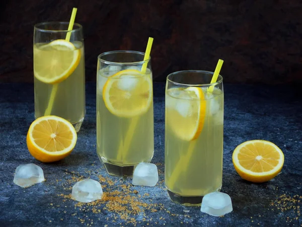 Drie glazen van koude zelfgemaakte limonade met citroen segmenten, ijsblokjes, bruine suiker en rietjes op donkere achtergrond. Kopiëren van ruimte — Stockfoto