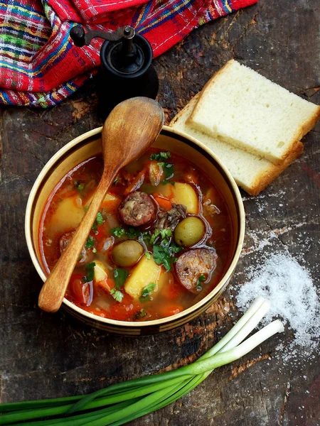 Hearty homemade soup with potatoes, carrots, sausages and olives in a clay bowl on a wooden background.