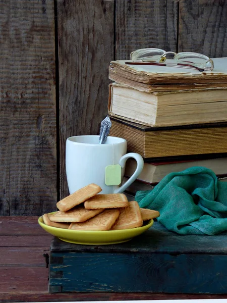 The composition of a stack of old books, tea cups, glasses and plates of sugar cookies on a wooden background. Vintage photo. Side view. copy space — Stock Photo, Image