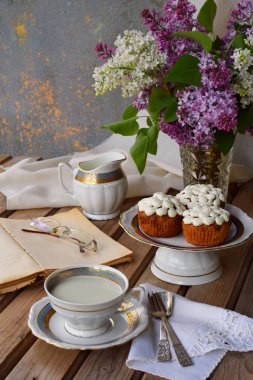Buket beyaz ve mor leylak, lor kremalı kek romantik bileşimi sütlü, eski kitap ahşap arka plan üzerinde çay bardağı.