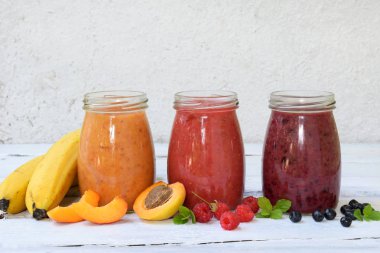 Turuncu, kırmızı ve mor smoothies taze malzemeler - muz, kayısı, ahududu ve hafif bir arka plan üzerinde yaban mersini. Sağlıklı gıda kavramı. Vejetaryen ürün. Organik çiğ içki. Kopya alanı.