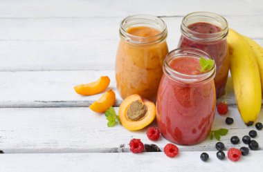 Turuncu, kırmızı ve mor smoothies taze malzemeler - muz, kayısı, ahududu ve hafif bir arka plan üzerinde yaban mersini. Sağlıklı gıda kavramı. Vejetaryen ürün. Organik çiğ içki. Kopya alanı.