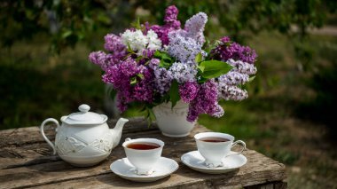 Ahşap arka planda vazoda leylak çiçekleri. Kahvaltı saatinde masada çay fincanı.