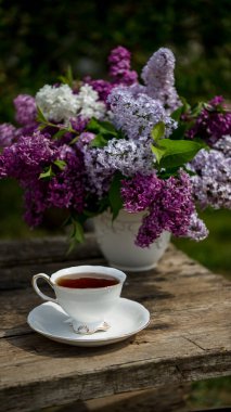 Ahşap arka planda vazoda leylak çiçekleri. Kahvaltı saatinde masada çay fincanı.
