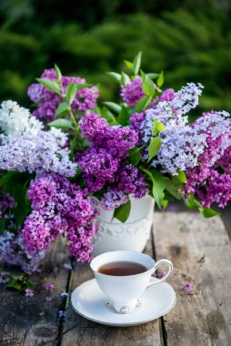 Ahşap arka planda vazoda leylak çiçekleri. Kahvaltı saatinde masada çay fincanı.