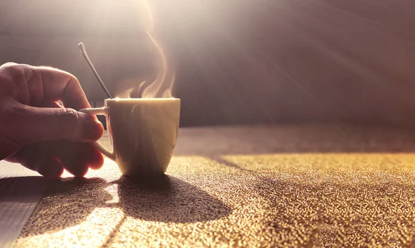 a steaming white ceramic cup containing coffee on a table lit by the light of the dawn sun. A male hand holds the cup by the handle. Morning coffee break. Relaxing moment. Back lit