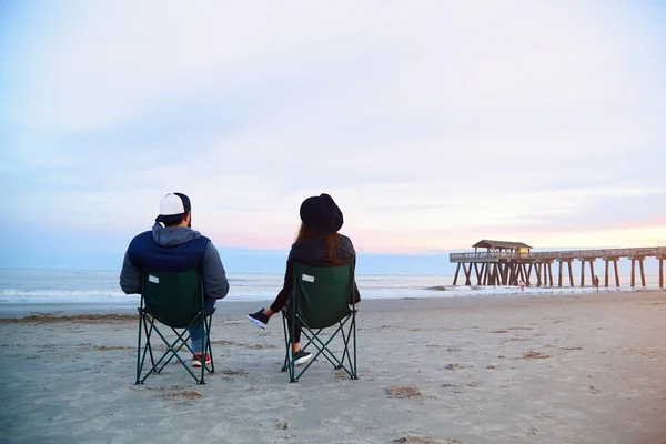 夫妻坐在甲板上的椅子上看美丽的日落海滩 免版税图库照片