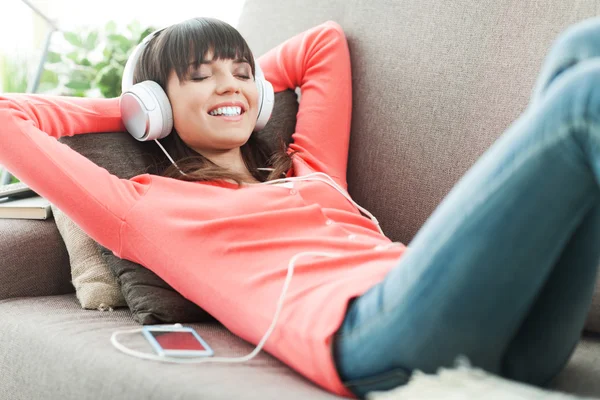 Ung attraktiv kvinde slapper af på sofaen derhjemme, hun lytter til musik med hovedtelefoner - Stock-foto