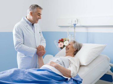 Doktor hastanedeki kıdemli bir hastayı ziyaret ediyor. Elini tutuyor, gülümsüyor ve onu rahatlatıyor.