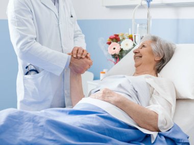 Doktor hastanede yaşlı bir hastayı ziyaret ediyor, elini tutuyor, gülümsüyor ve onu rahatlatıyor.