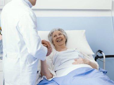 Doktor hastanedeki kıdemli bir hastayı ziyaret ediyor. Elini tutuyor, gülümsüyor ve onu rahatlatıyor.