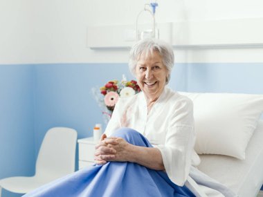 Gülümseyen yaşlı kadın hastane yatağında poz veriyor ve kameraya, sağlık hizmetlerine ve yaşlı bakım konseptine bakıyor.