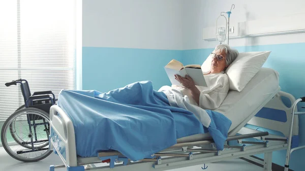 老妇人躺在医院的床上看书 — 图库照片