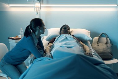 Hastanede genç arkadaşına kanser konusunda yardım eden ilgili bir kadın, yatağının yanında oturuyor ve onu kontrol ediyor.