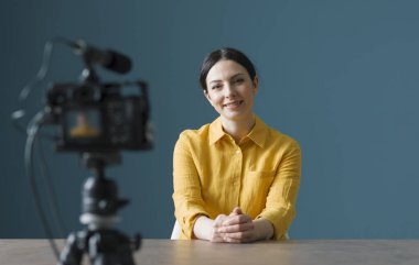 Gülümseyen kadın kameranın önünde oturmuş video blogu yazıyor.