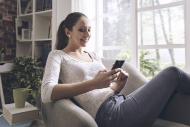 Evde akıllı telefon kullanan mutlu genç kız, bir pencerenin yanındaki koltukta dinleniyor.