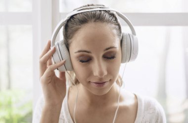 Mutlu rahat kadın evde müzik kulaklık kullanarak, bir pencere yanında oturuyor