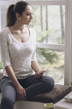 Pencerenin kenarında oturan ve akıllı telefonuyla iletişim kuran genç kız sohbet ediyor ve dinleniyor.