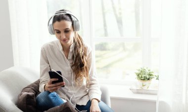 Oturma odasındaki koltukta oturan ve kulaklık ve akıllı telefon kullanarak müzik dinleyen genç bir kadın.