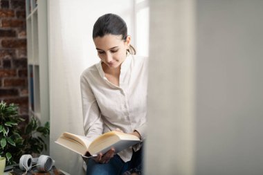 Genç, dingin bir kadın evde dinleniyor ve pencerenin yanında kitap okuyor.