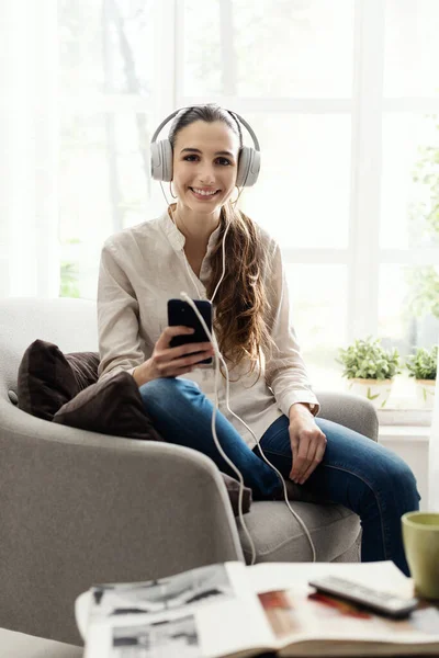 居間の椅子に座ってヘッドフォンやスマートフォンを使って音楽を聴いている若い笑顔の女性 — ストック写真