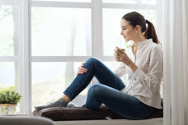 窓の横の家でリラックスしてコーヒーを飲んでいる若い女性 — ストック写真