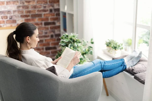 年轻的女人在家里靠窗休息 她坐在扶手椅上看书 — 图库照片