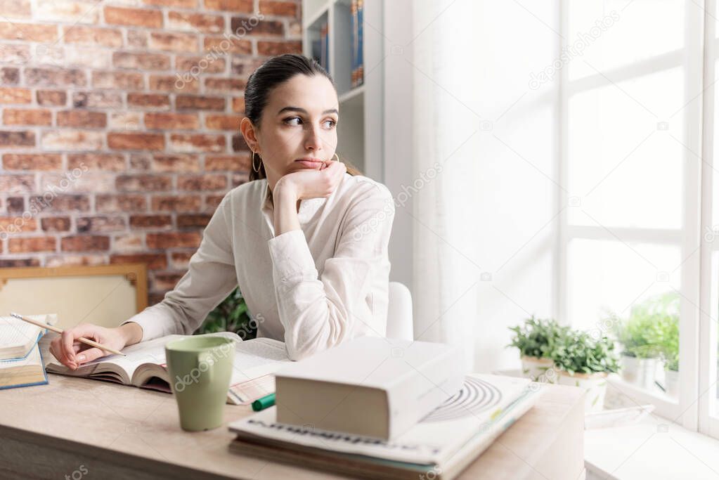 Young woman sitting at desk and studying at home, she is looking away and thinking
