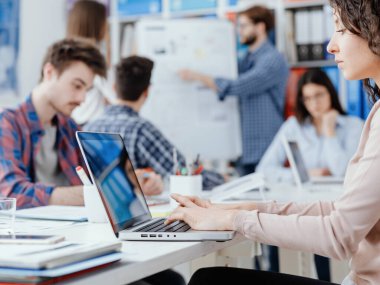 Genç iş adamları ofiste toplanıyor ve çalışıyor, bir kadın ön planda dizüstü bilgisayar kullanıyor: başlangıç ve yenilik kavramı