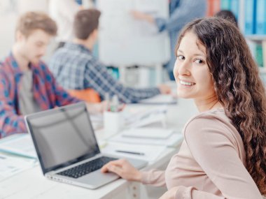 Bir grup üniversite öğrencisi birlikte okuyorlar. Bir kız öğrenci gülümsüyor ve ön planda dizüstü bilgisayar kullanıyor.