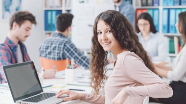 Bir grup üniversite öğrencisi birlikte okuyorlar. Bir kız öğrenci gülümsüyor ve ön planda dizüstü bilgisayar kullanıyor.