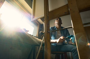 Stüdyosunda oturan ve tablosu üzerinde çalışan yaratıcı genç bir sanatçı: elinde fırça ve boya dolu bir kavanozla oturuyor.