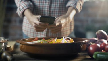 Kadın taze sebzelerle sağlıklı bir yemek hazırlıyor ve akıllı telefonuyla fotoğraf çekiyor.