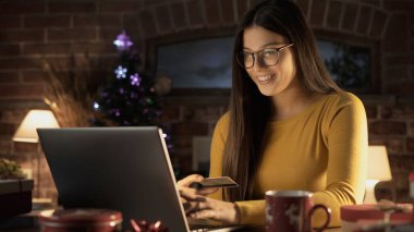Bilgisayarına bağlanan ve Noel hediyelerini kredi kartıyla, online alışveriş ve tatil konseptiyle alan mutlu genç bir kadın.