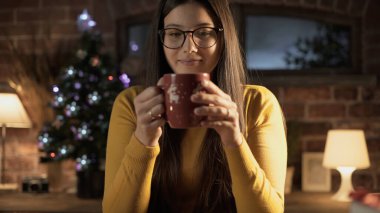 Mutlu genç bir kadının evde çay içmesi, arka planda Noel ağacı, tatil konsepti.