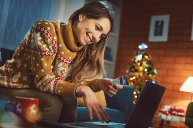 Mutlu kadın evdeki kanepede dinleniyor ve online alışveriş yapıyor, arka planda Noel ağacı var.