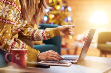 Noel 'de internetten alışveriş yapan bir kadın: Evdeki koltukta oturuyor, bilgisayarına bağlanıyor ve elinde bir kredi kartı, arka planda Noel ağacı tutuyor.