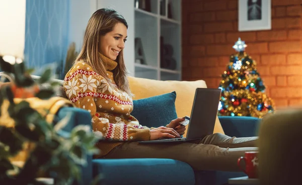 幸せな若い女性は自宅でソファに座って クリスマスプレゼントのためにオンラインショッピングをして 彼女は彼女のラップトップと接続し クレジットカードを保持しています — ストック写真