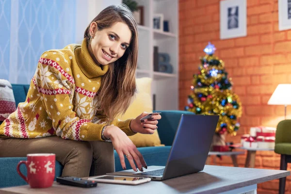快乐的女人坐在沙发上 用笔记本电脑在网上购物 拿着信用卡 对着相机笑着 — 图库照片