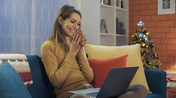 一位快乐的年轻女士坐在家里的沙发上 与笔记本电脑相连 她在网上收到了一个美丽的惊喜 背景是圣诞树 — 图库照片