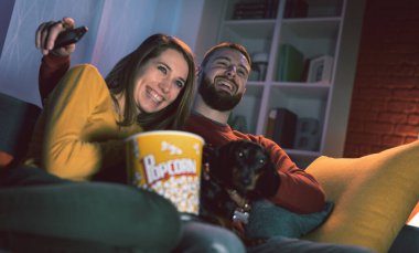 Mutlu çift evde köpekleriyle kanepede dinleniyor: TV şovları izliyor, patlamış mısır yiyor ve birlikte gülüyorlar.