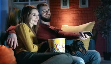 Mutlu çift evlerindeki kanepede köpekleriyle dinleniyor, televizyon seyrediyor ve patlamış mısır yiyor, uzaktan kumandayı elinde tutuyor.