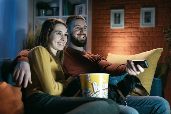 一对快乐的夫妇和他们的狗在沙发上休息 他们正在看电视节目 吃爆米花 他拿着遥控器 — 图库照片