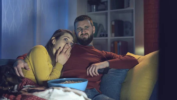 若いですカップル座っていますザカウチに自宅でホラー映画を一緒にテレビで見て 彼は彼のガールフレンドを抱きしめています — ストック写真