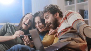 Mutlu arkadaşlar evdeki kanepede oturuyor ve akıllı telefonları, birliktelikleri ve teknoloji konseptiyle sosyal iletişim kuruyorlar.