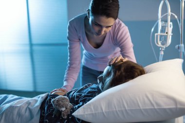 Genç bir anne, küçük çocuğuna gece hastanede yatarken yardım ediyor. Endişeli ve başını okşuyor.