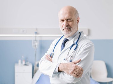 Profesyonel doktor hastane yatağının yanında duruyor ve katlanmış kollar, sağlık uzmanları ve tıp konseptiyle poz veriyor.