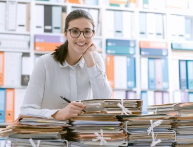 Gülümseyen genç ofis çalışanı kağıt yığınlarına yaslanıyor.