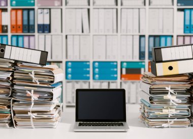 Ofiste yığınla evrak ve masaüstünde dizüstü bilgisayar.