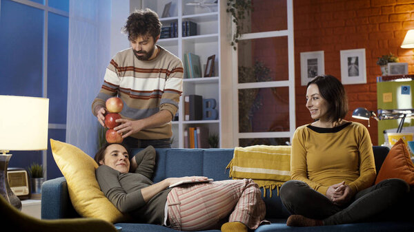 Young friends spending time together at home, the man is stacking apples on his friends head while she is lying on the sofa